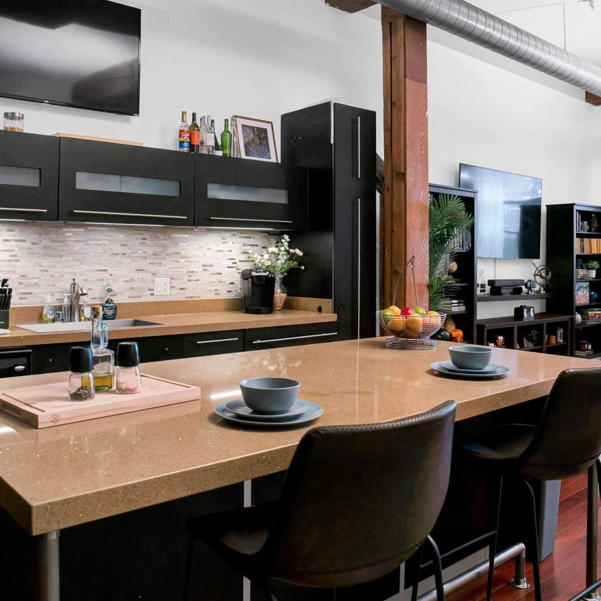 A modern kitchen with a central island, equipped with two stools, a stovetop, and a variety of kitchen appliances, against a backdrop of exposed wooden beams.