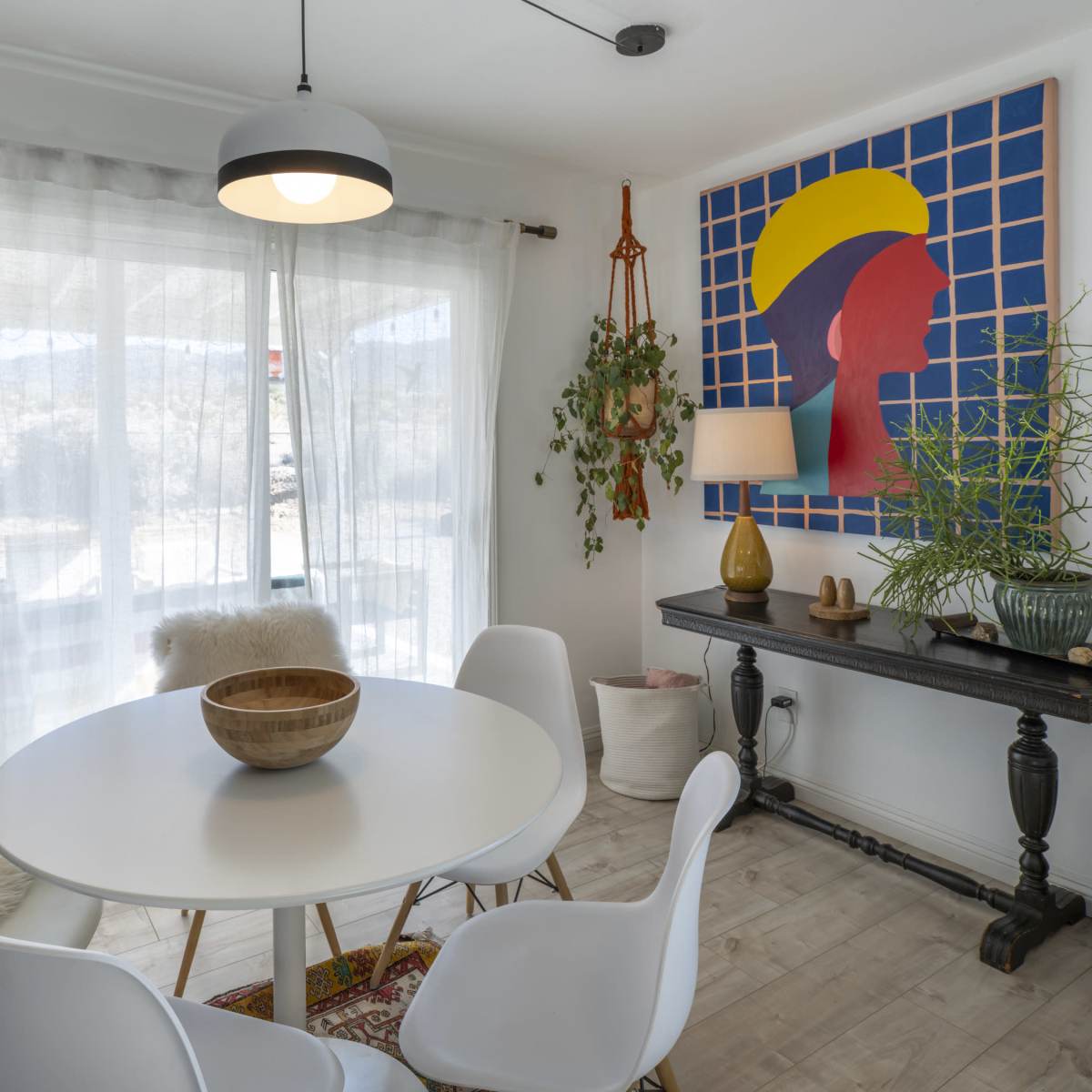 A modern dining area features a round white table surrounded by four chairs, with a large artwork on the wall and a window allowing natural light to enter.