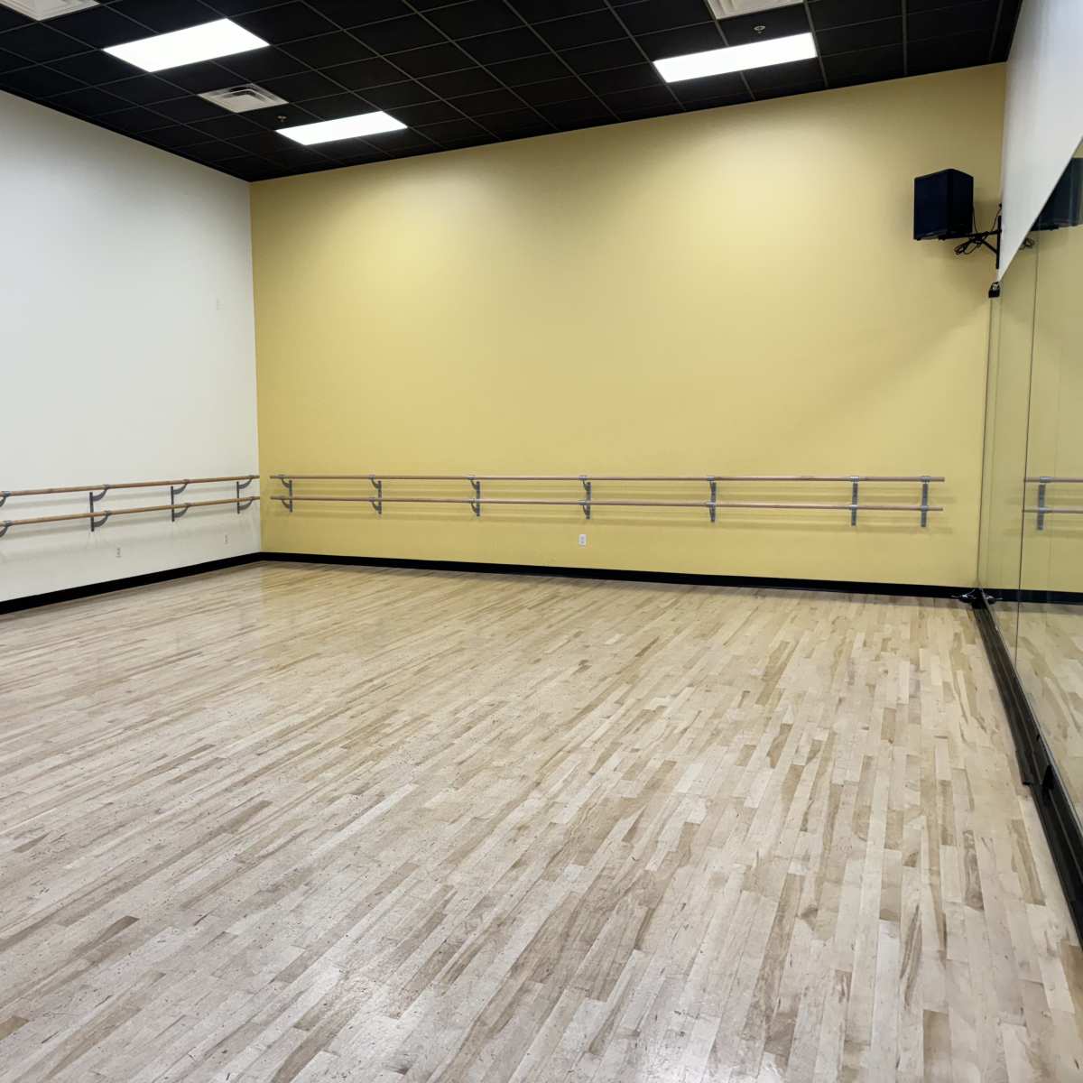 An empty dance studio with wooden flooring, yellow walls, and mirrored panels along one side.