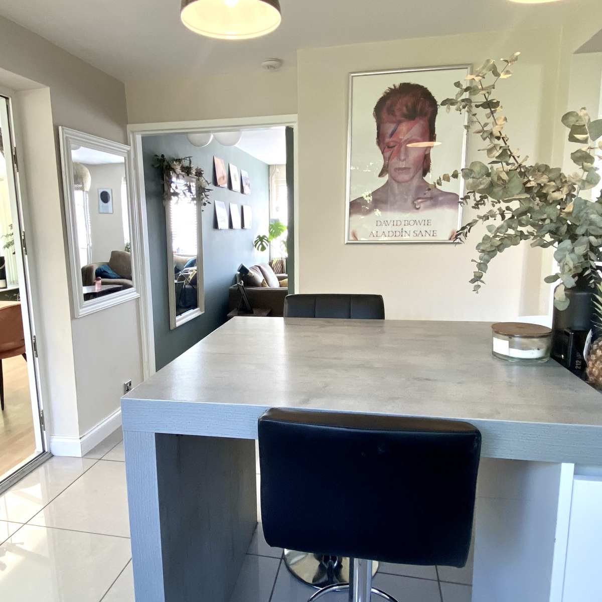 A modern kitchen area with a bar-style countertop, a poster of David Bowie on the wall, and a view of a sunlit room with seating in the background.