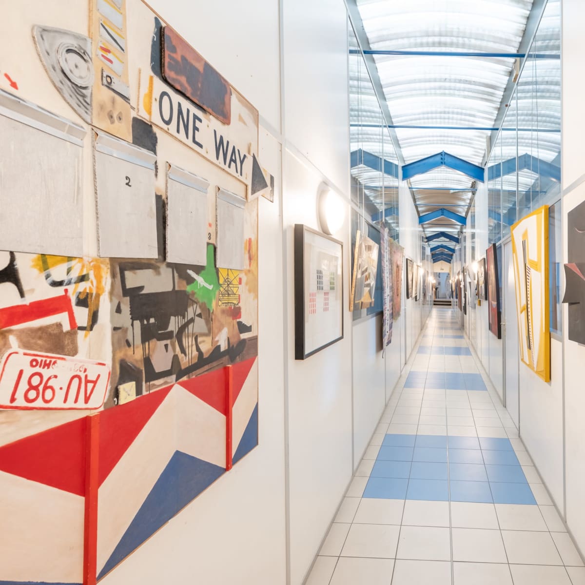 A long corridor lined with various modern art pieces on the walls, with a glass ceiling allowing natural light to illuminate the space.