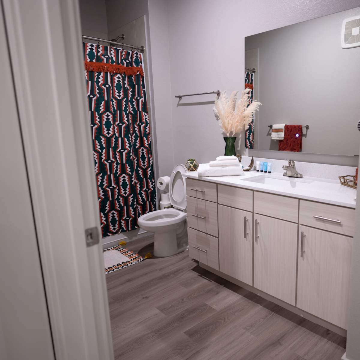 A modern bathroom features a white countertop, a mirror, a toilet, and a shower curtain with a colorful geometric pattern.