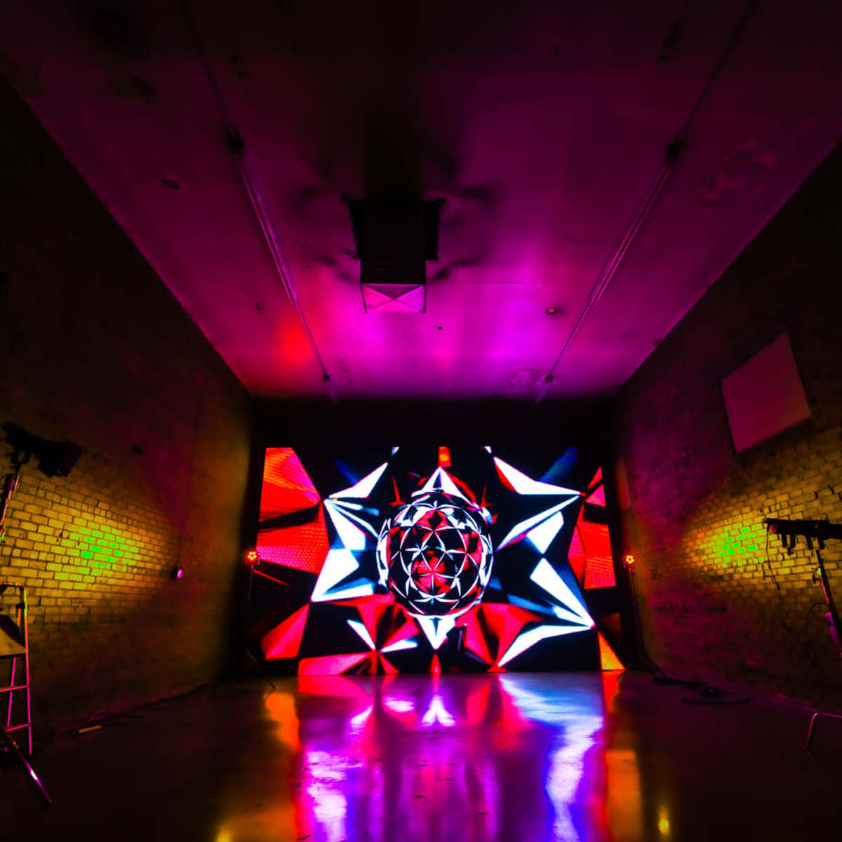 A dimly lit room with a vibrant, colorful light display featuring geometric patterns on a rear wall and stage lighting equipment set up in the corners.