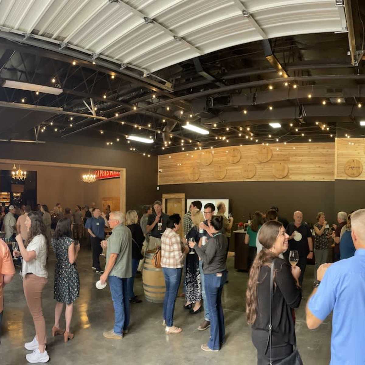 A crowd of people mingles inside a spacious venue featuring wooden accents and decorative lighting.