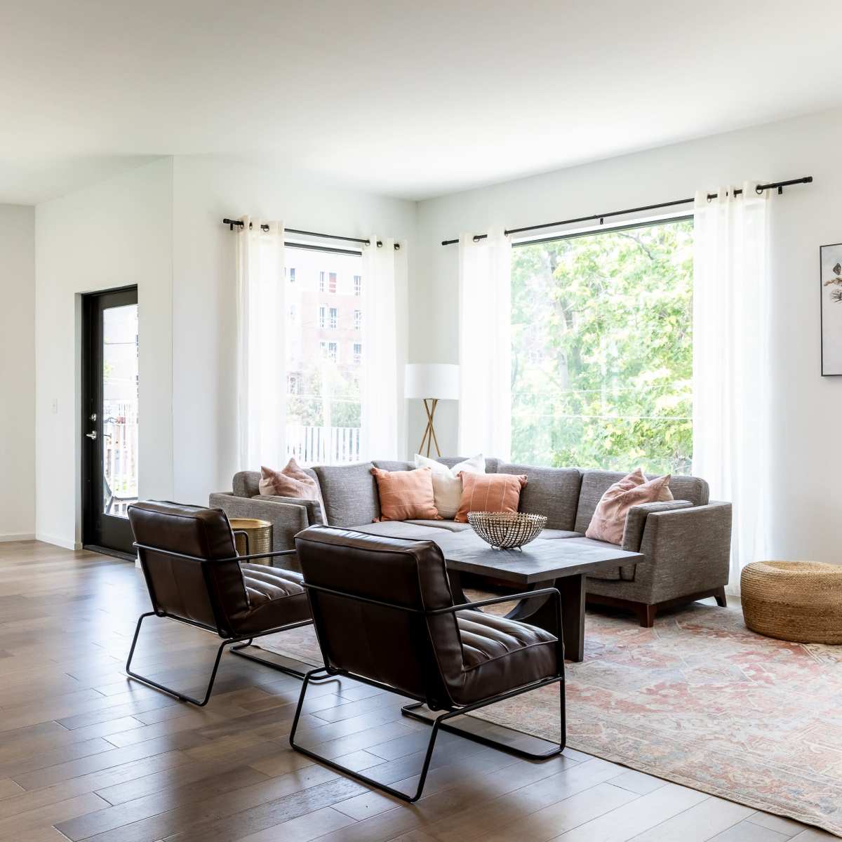 A bright, open living space featuring a seating area with a gray sofa and black chairs, a dining table for four, and large windows letting in natural light.