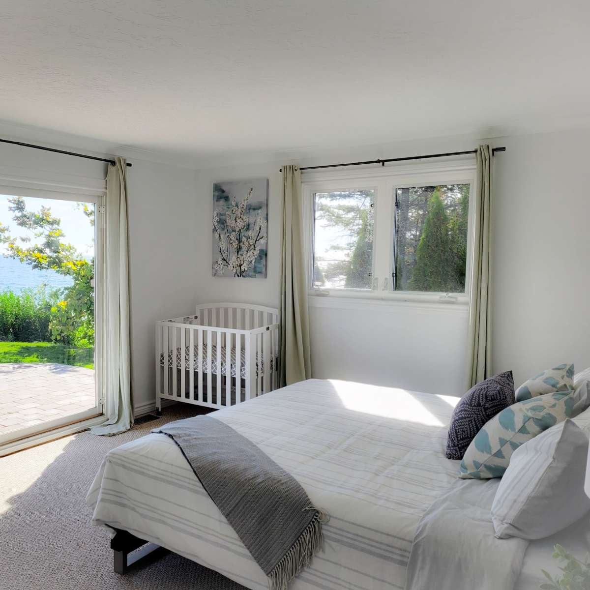 A bright bedroom features a crib next to a large window overlooking a view of the ocean and lush greenery outside.