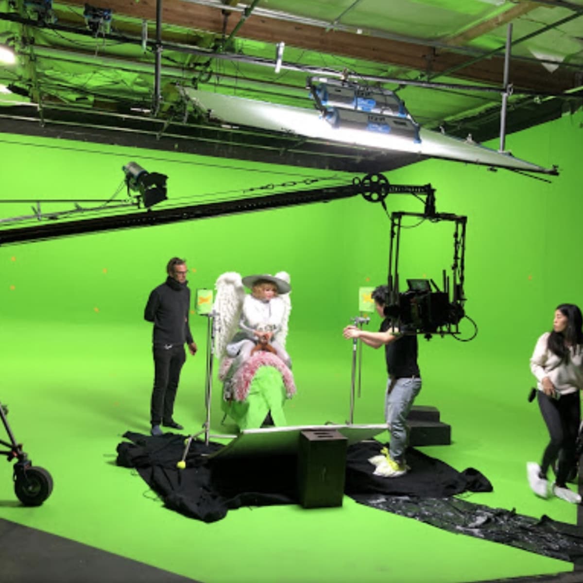 A production crew sets up a green screen studio with a cameraman filming a person dressed in costume seated on a large chair.