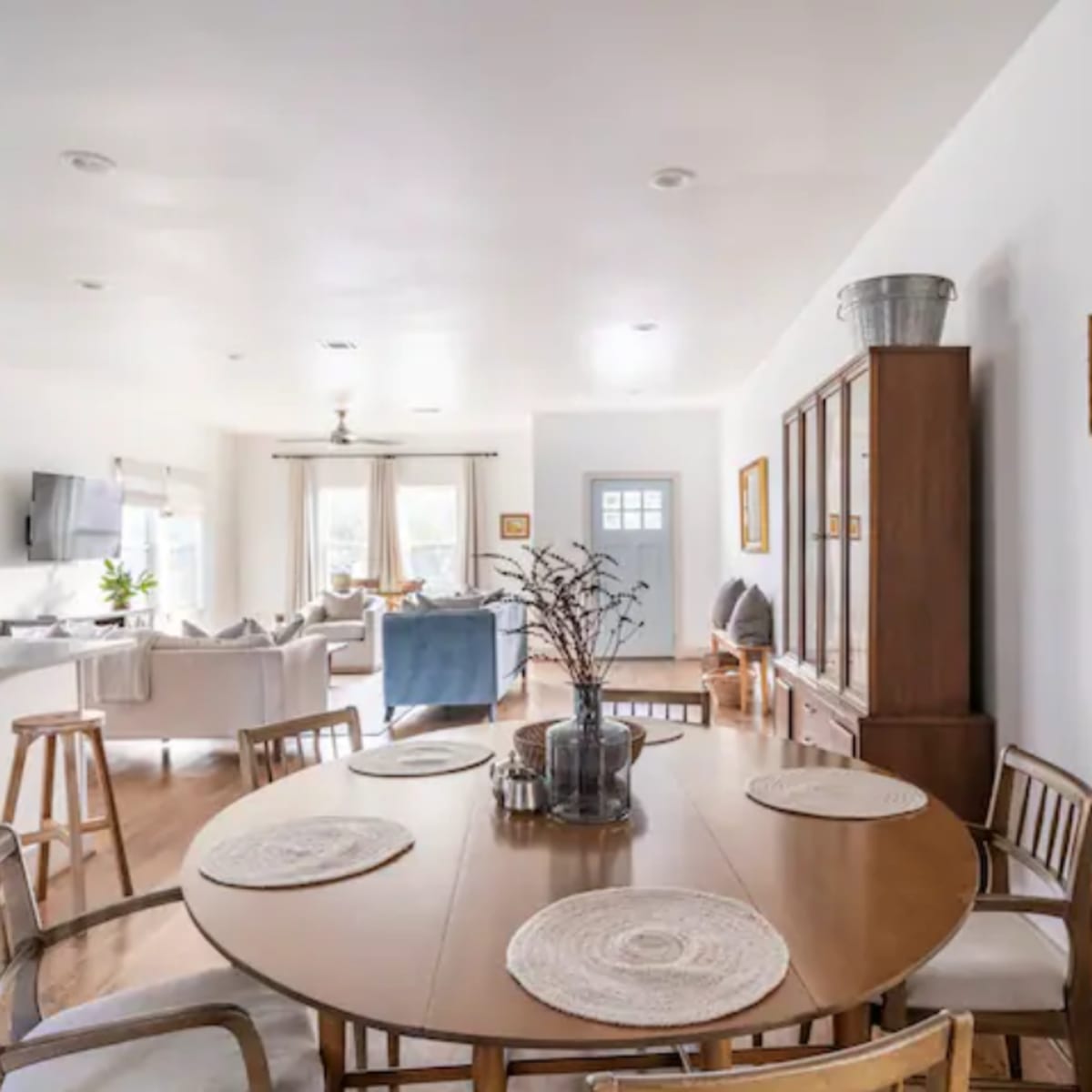 A dining area features a round wooden table surrounded by chairs, with a view of a bright living space beyond.