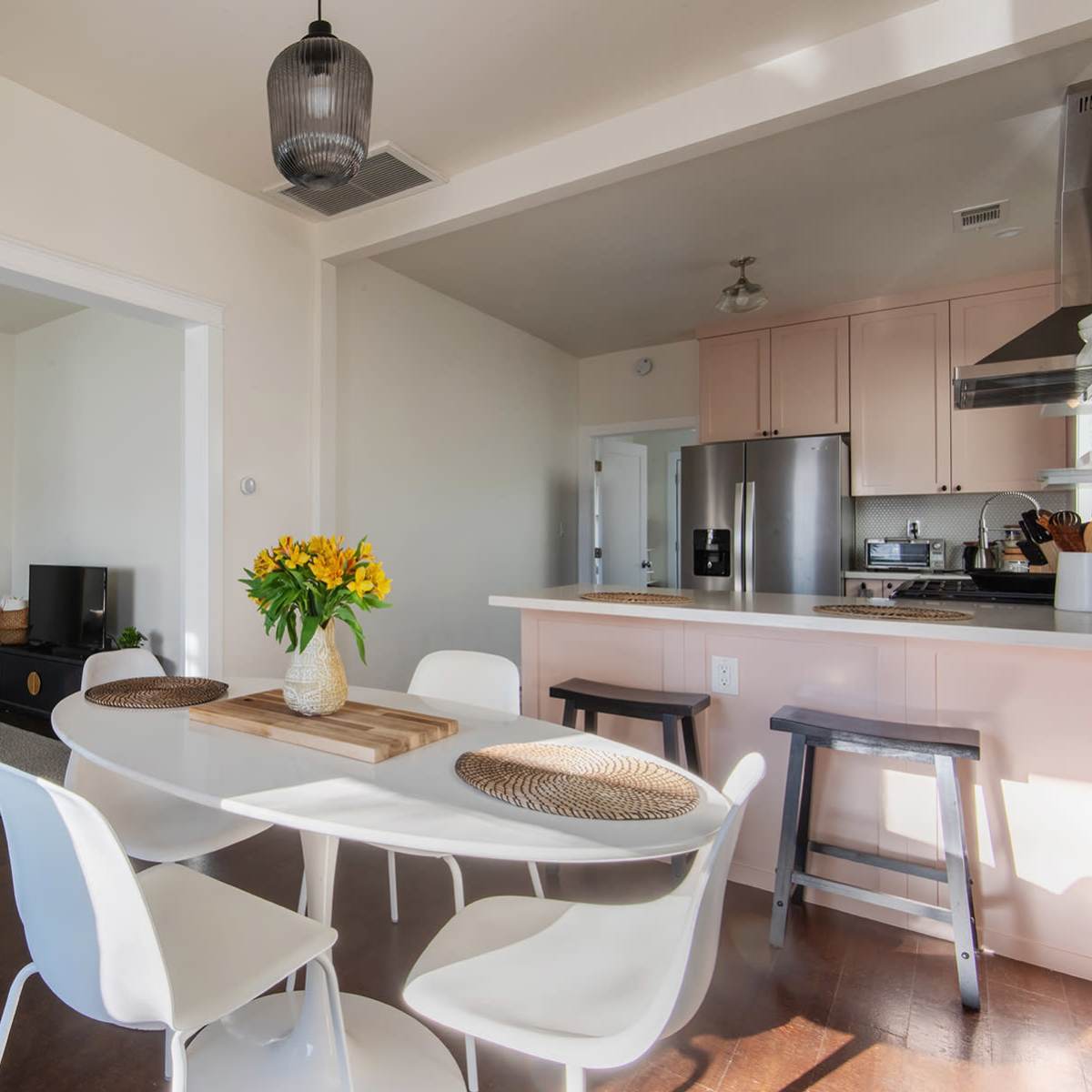 A modern dining area with a round table, white chairs, and a vase of flowers, adjacent to a living space featuring a mirror, a television, and a light-colored sofa.