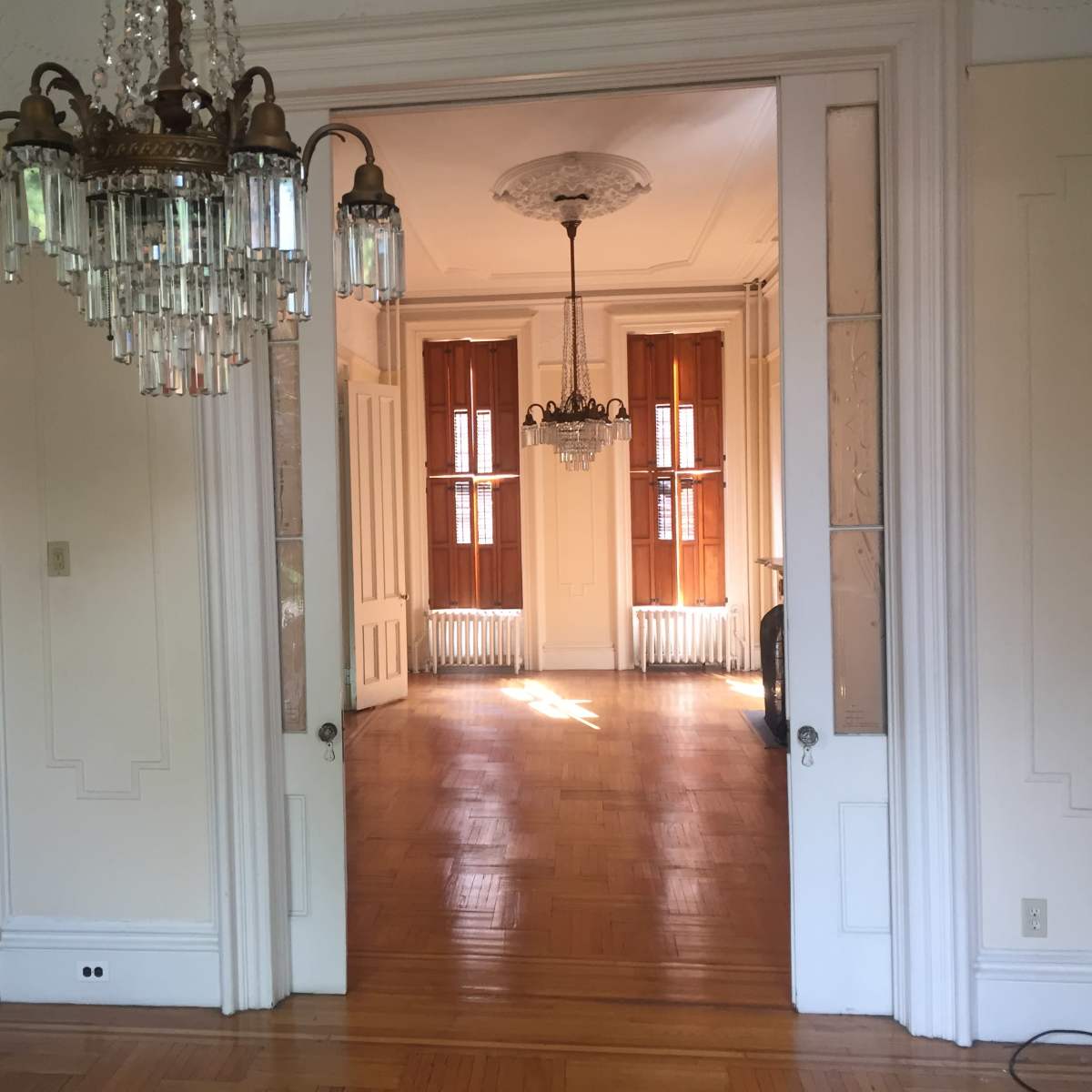 A well-lit room with a chandelier and another room visible through an open doorway, featuring large windows and hardwood floors.