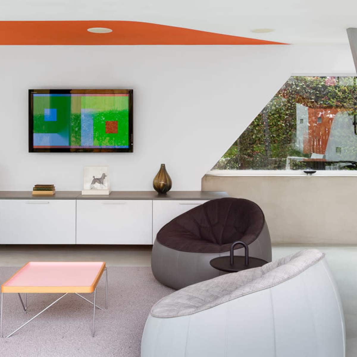 The room features modern furniture, a large window with greenery outside, and a staircase with a support beam.