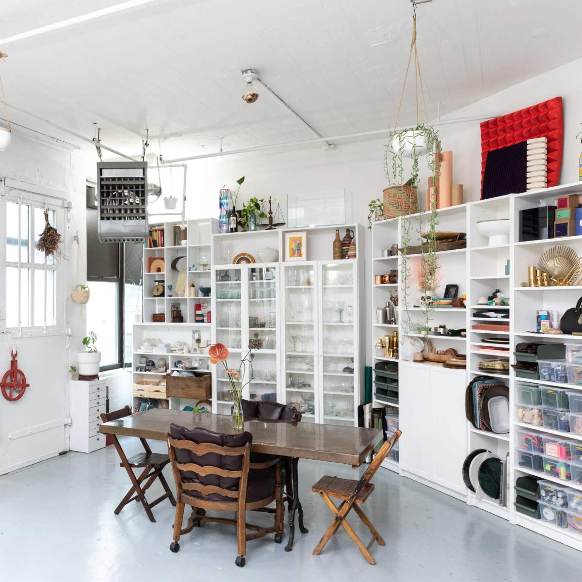 A bright room featuring a wooden dining table surrounded by chairs, with white shelves filled with various items and plants, and large windows allowing natural light to enter.
