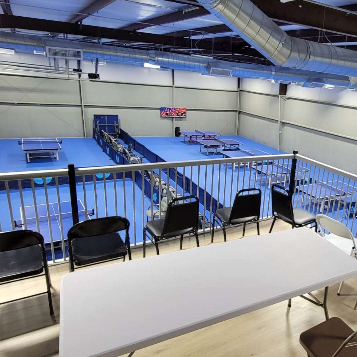 An indoor table tennis facility with blue floors, multiple tables arranged for play, and a viewing area overlooking the courts.