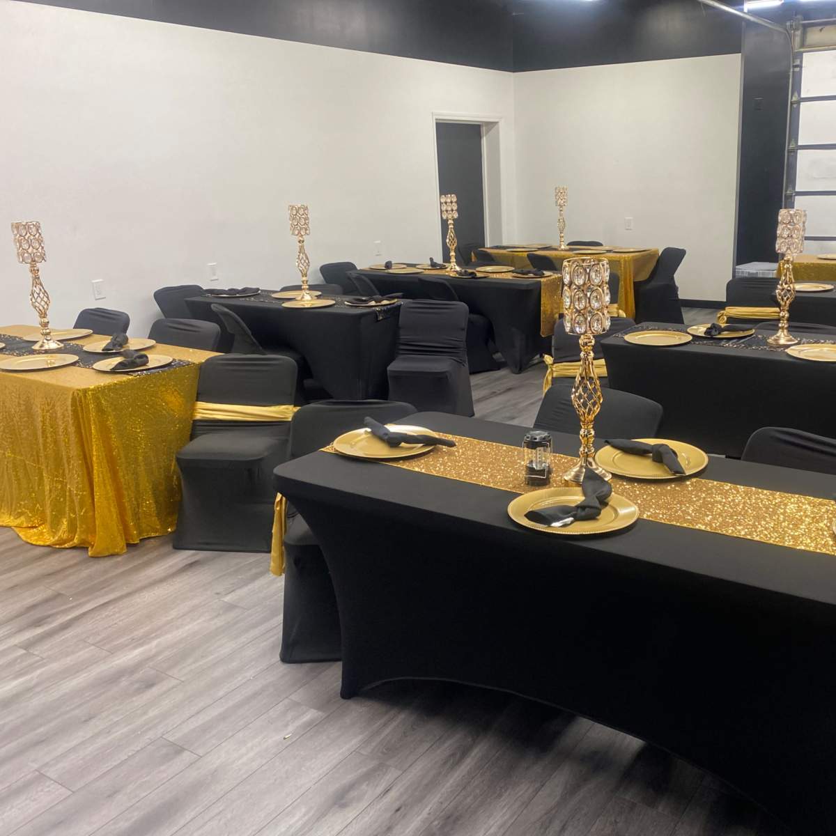 A banquet setup with several tables covered in black and gold tablecloths, each adorned with decorative centerpieces and place settings.