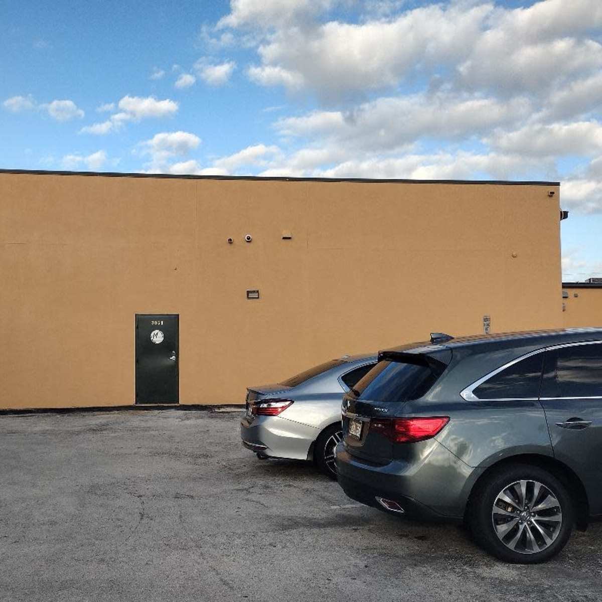 A yellow building wall with a green door, surrounded by parked cars in a lot under a partly cloudy sky.