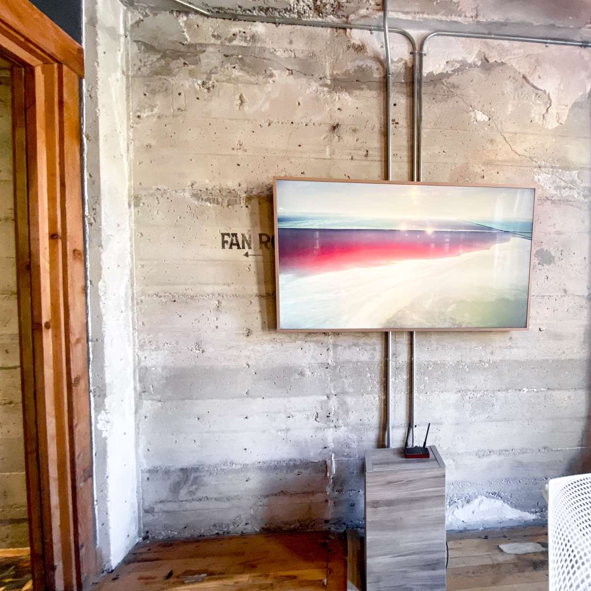 A framed artwork is mounted on a concrete wall, with visible electrical conduits running alongside it, and a small wooden pedestal next to a white chair.