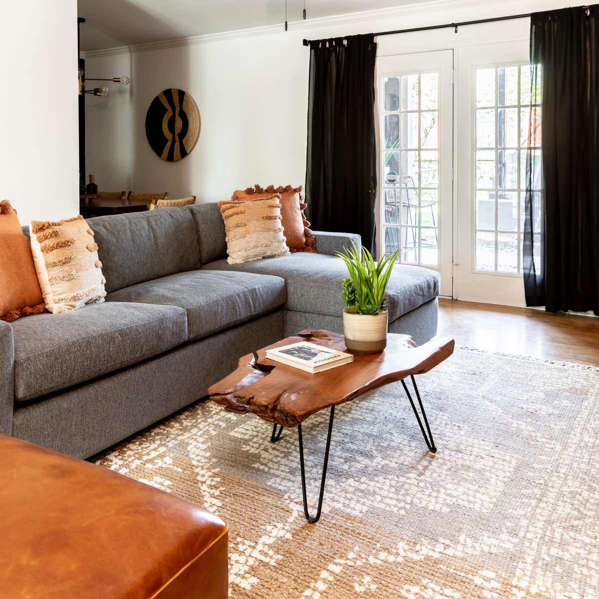 A modern living room features a gray sectional sofa, a wooden coffee table, and large windows covered with black curtains.