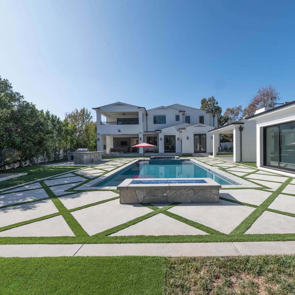 A modern two-story house with a swimming pool is surrounded by neatly patterned concrete and artificial grass landscaping.