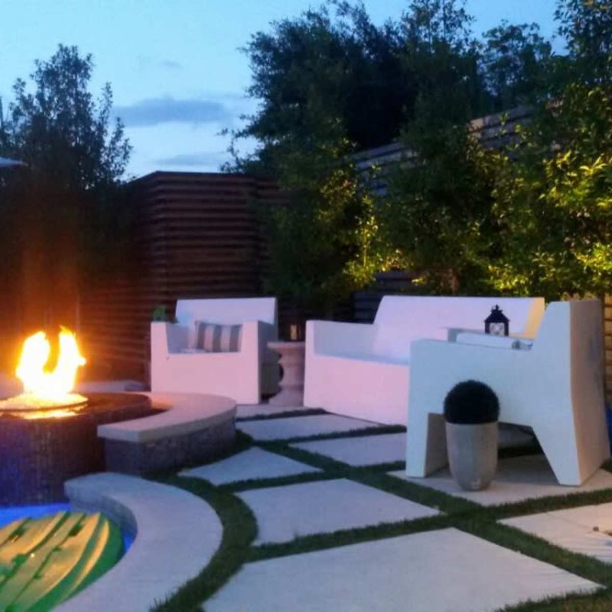 The scene features a cozy outdoor seating area beside a glowing fire pit, surrounded by greenery and soft lighting.