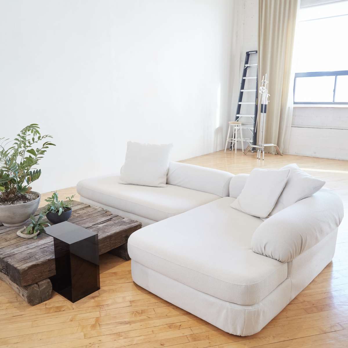 A light-colored sectional sofa and a rustic wooden coffee table are arranged in a spacious room with large windows and minimal furnishings.