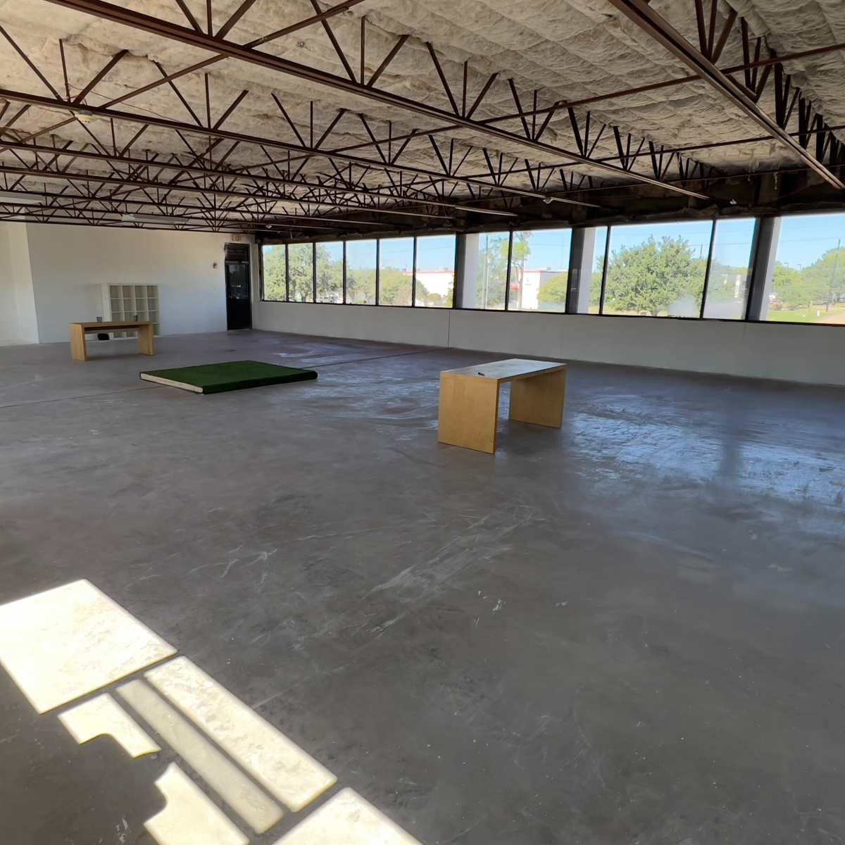 A spacious, empty room with large windows, a concrete floor, a few pieces of furniture, and exposed metal beams.