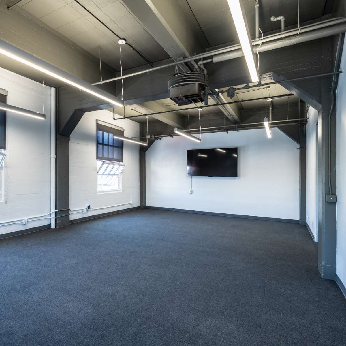 An empty room with gray carpet, featuring two windows, exposed beams, and a wall-mounted television.