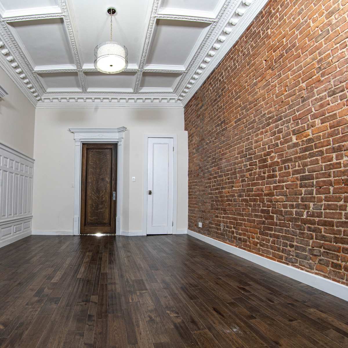 A spacious room featuring a mix of white paneling and a brick wall, with a wooden floor and a decorative ceiling.