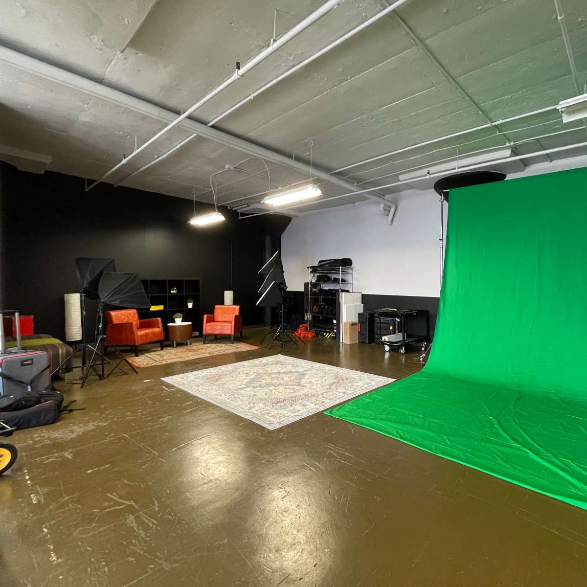 A spacious studio setting featuring a green screen, two orange chairs, a rug, and various photography equipment.