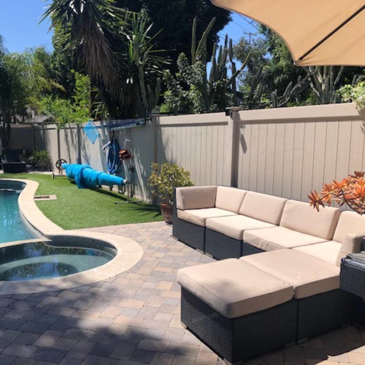 A backyard scene features a swimming pool, a seating area with sectional sofas, and a clothesline with blue fabric, surrounded by greenery and palm trees.
