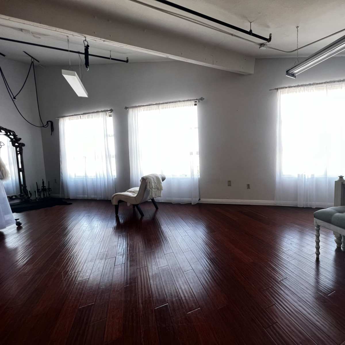 A spacious room with hardwood floors, two large windows covered by sheer curtains, and minimal furniture including a bed and a chair.