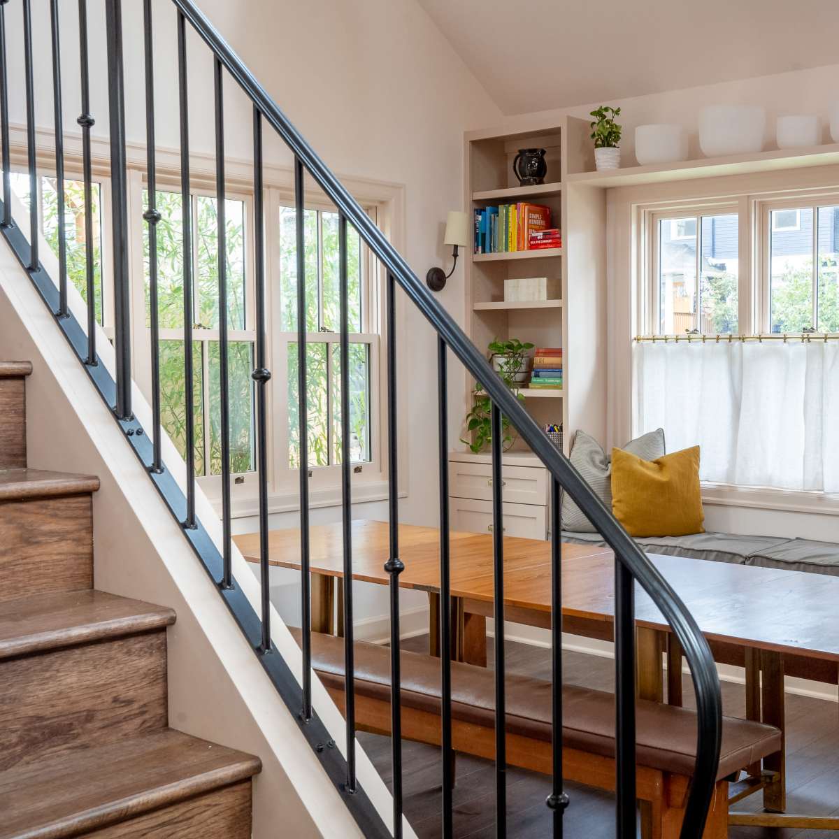 A well-lit interior space featuring a stairway, a dining table, and a cozy window seat surrounded by shelves and decorative items.