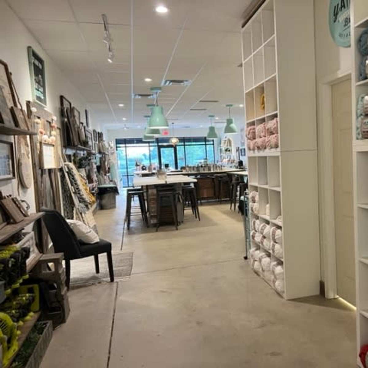 A well-lit craft store aisle lined with shelves of yarn and art supplies, leading towards a seating area with tables and chairs.