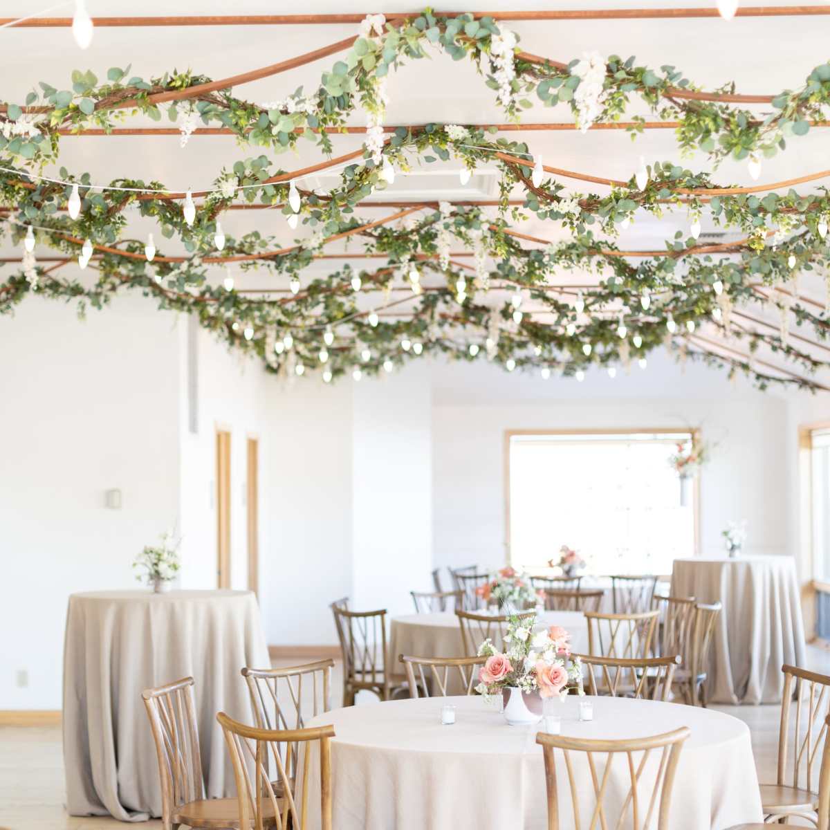 A banquet hall decorated with greenery and string lights, featuring round tables covered with light fabric.