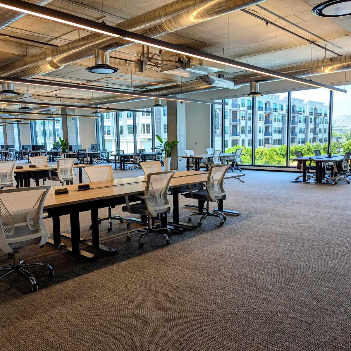 A spacious modern office with several rows of desks and chairs, large windows offering a view of buildings and mountains outside.