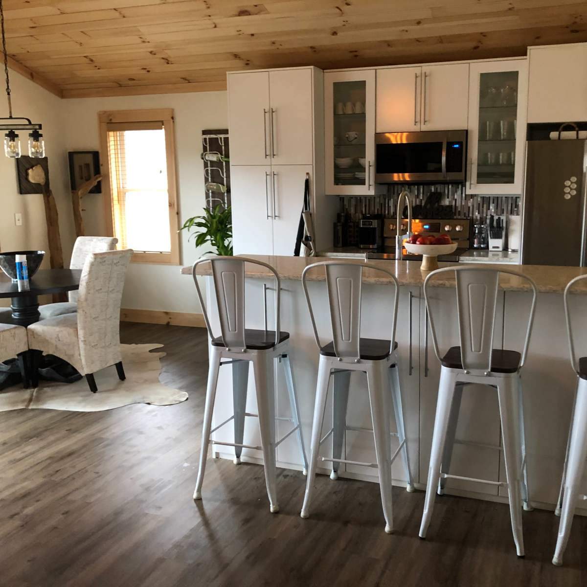 A modern kitchen with a bar area featuring four metal stools, a dining table in the background, and wooden ceiling beams.