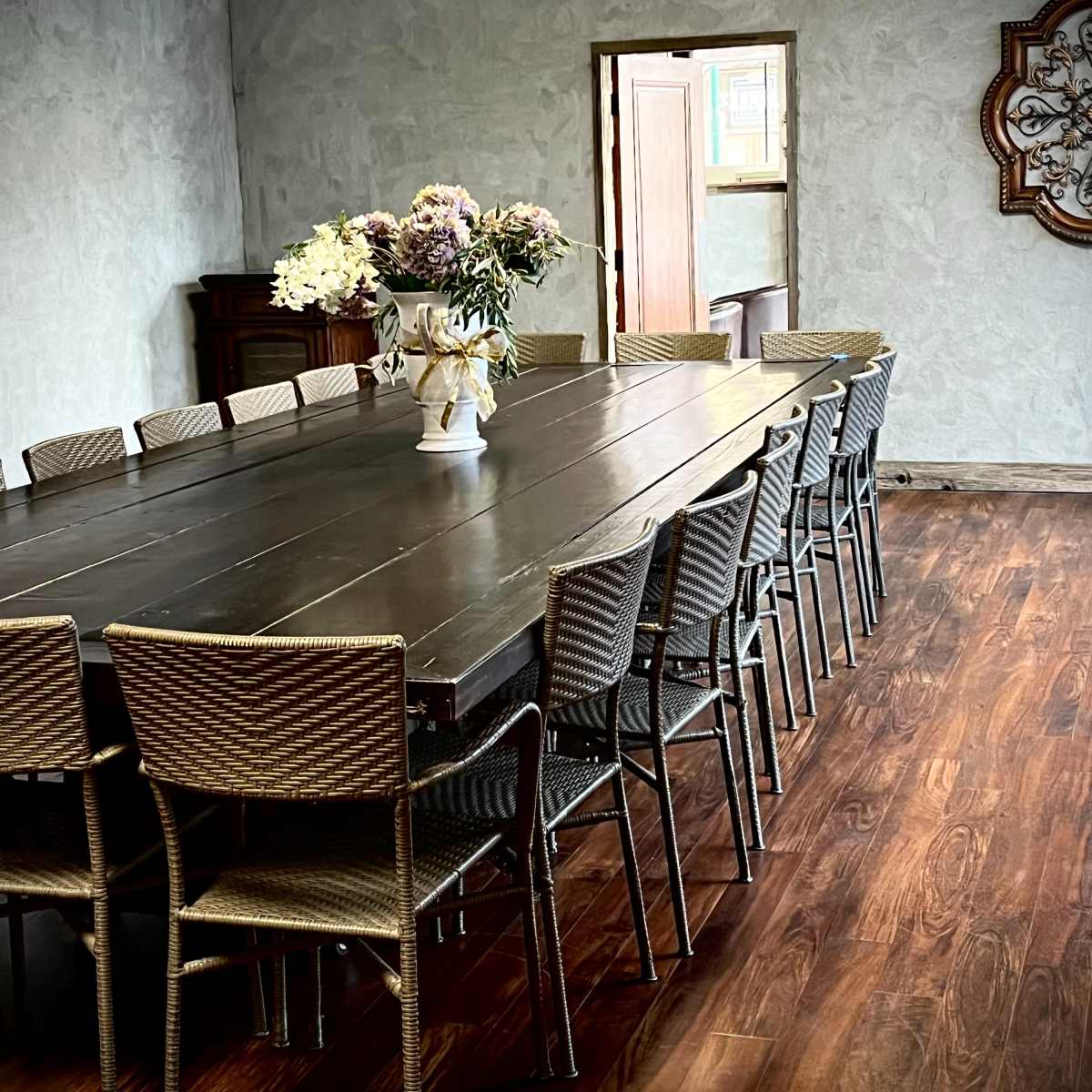 A large wooden table is surrounded by wicker chairs in a room with textured gray walls and a doorway in the background.