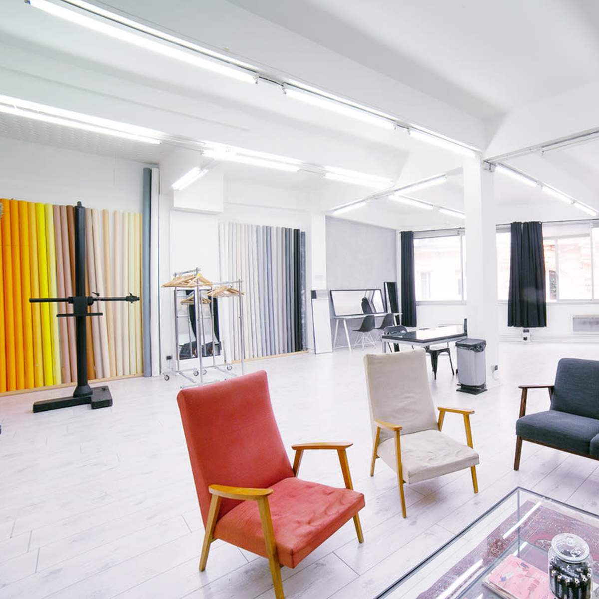 A bright, spacious studio featuring a sofa, two armchairs, and several backdrop stands against a large window.
