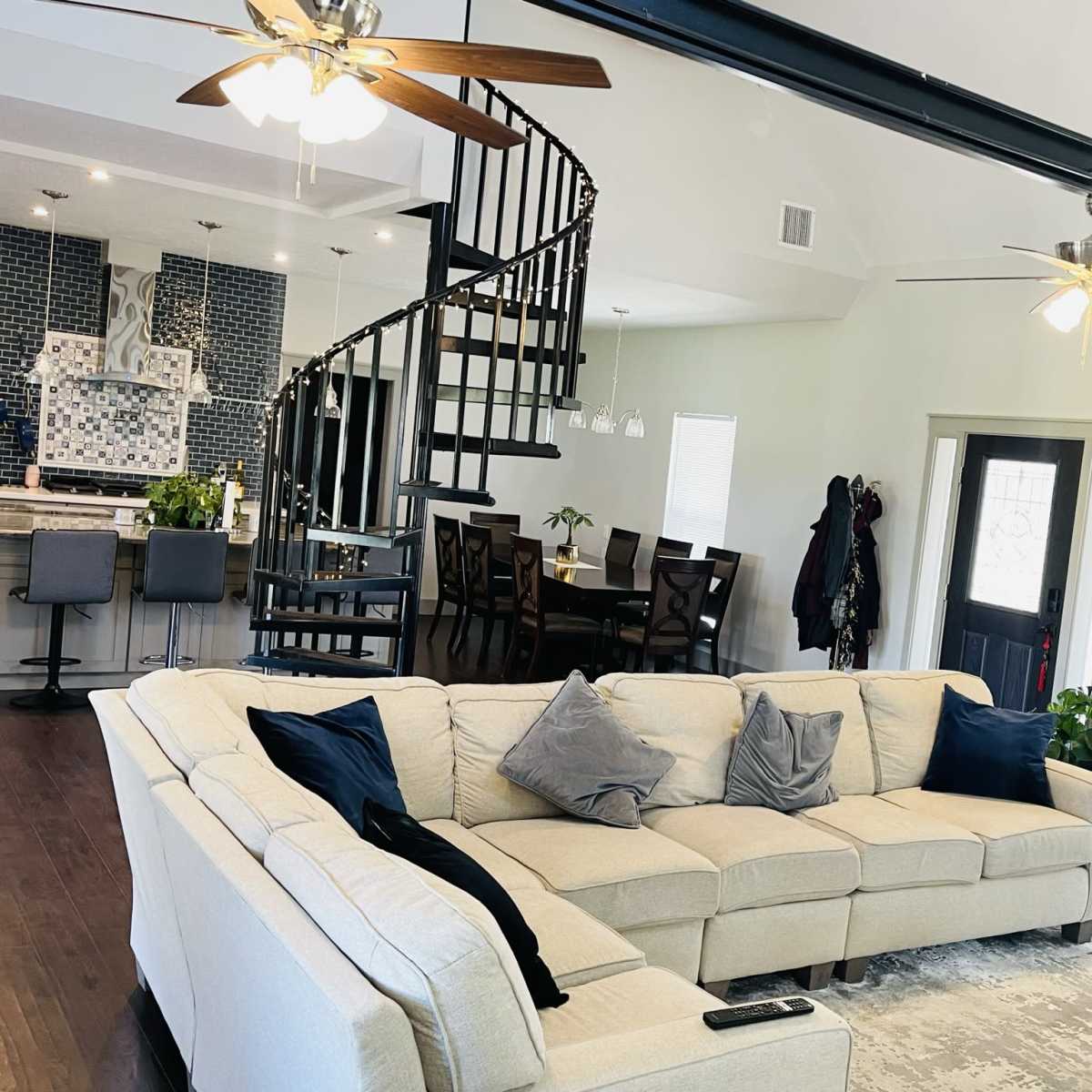 A spacious living room with a beige sectional sofa, a spiral staircase leading to an upper level, and a dining area in the background.