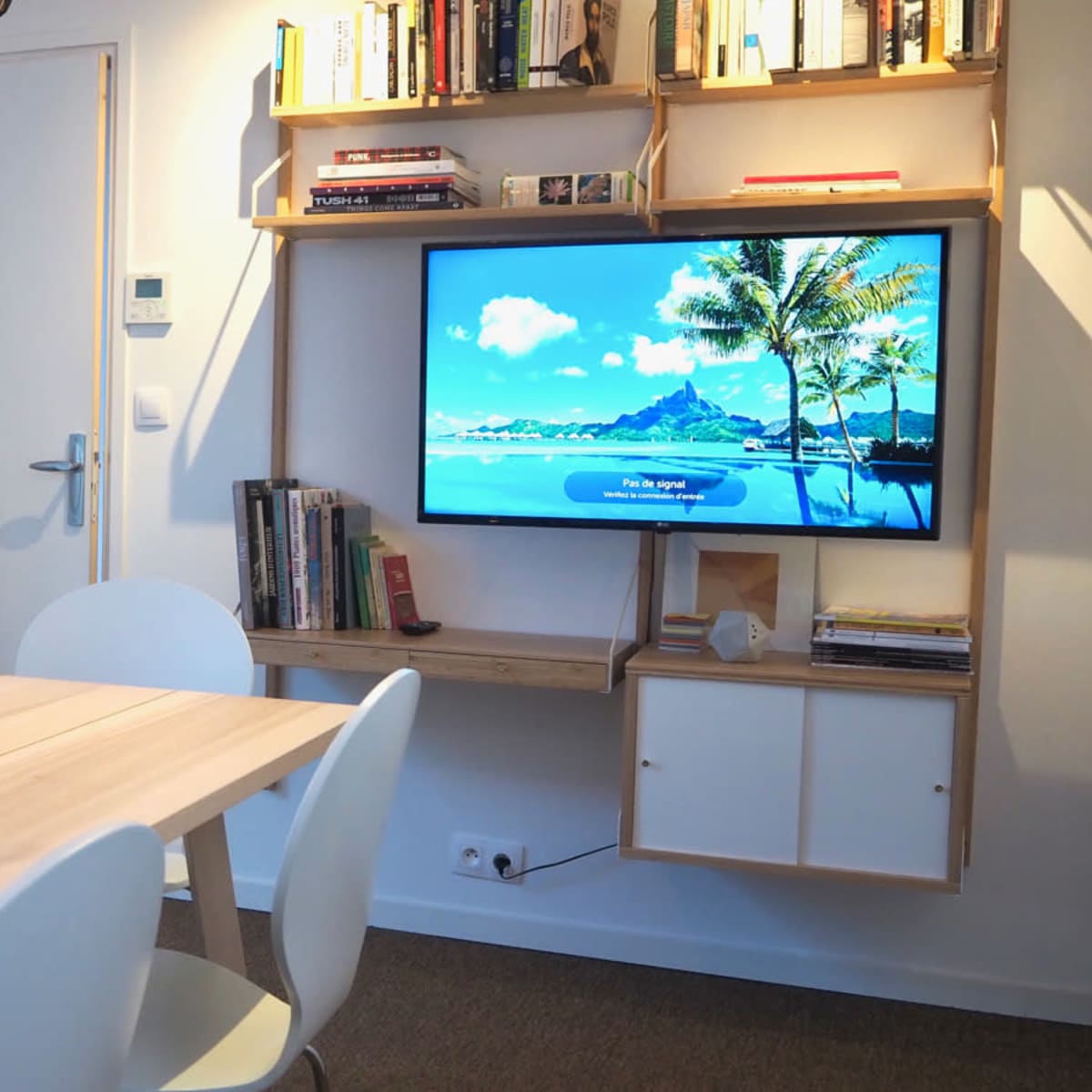 A well-lit modern living space features a wooden table, a wall-mounted TV displaying a beach scene, and a bookshelf filled with books and decorative items.