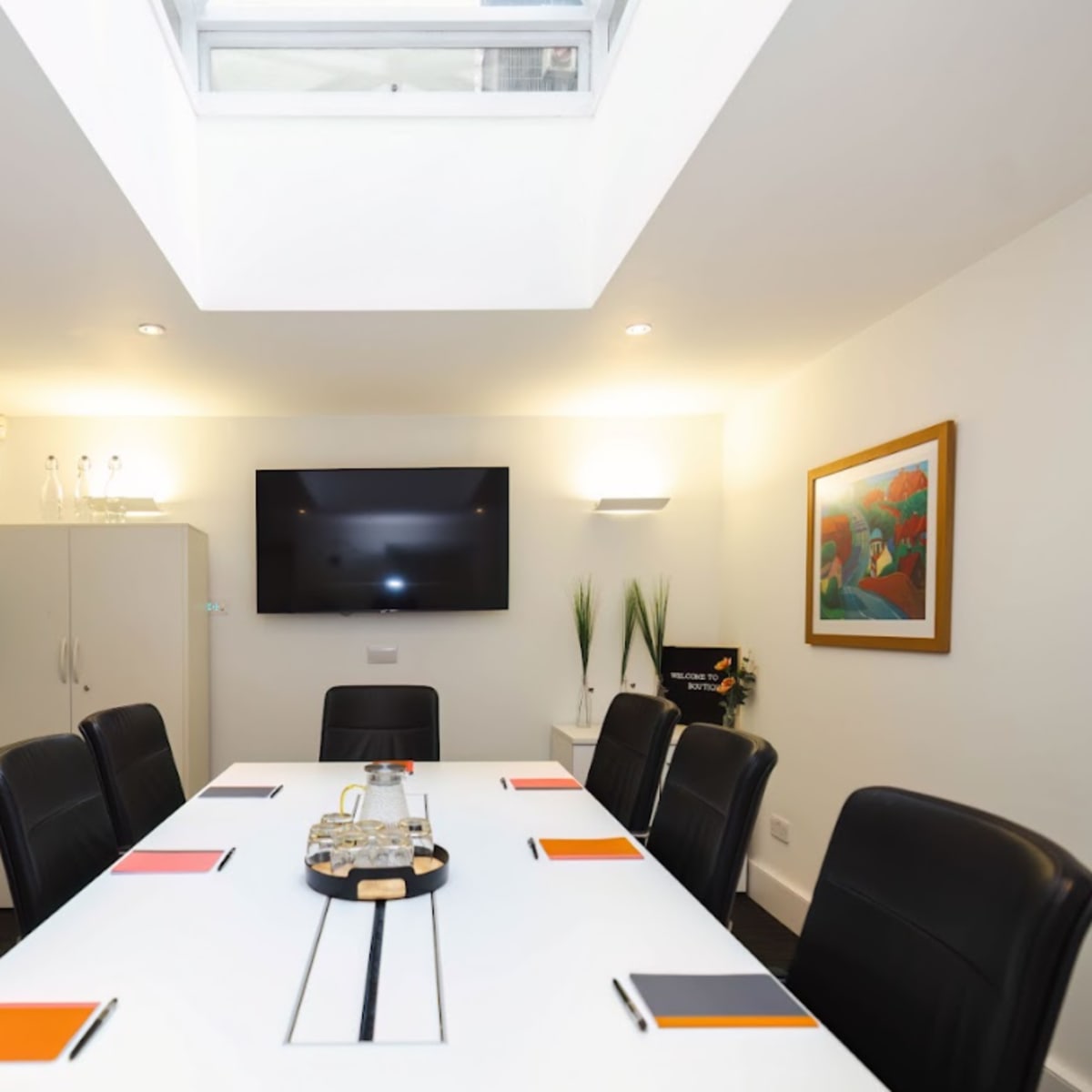 A modern conference room features a long white table surrounded by black leather chairs, with framed artwork on the walls and a television mounted on one wall.