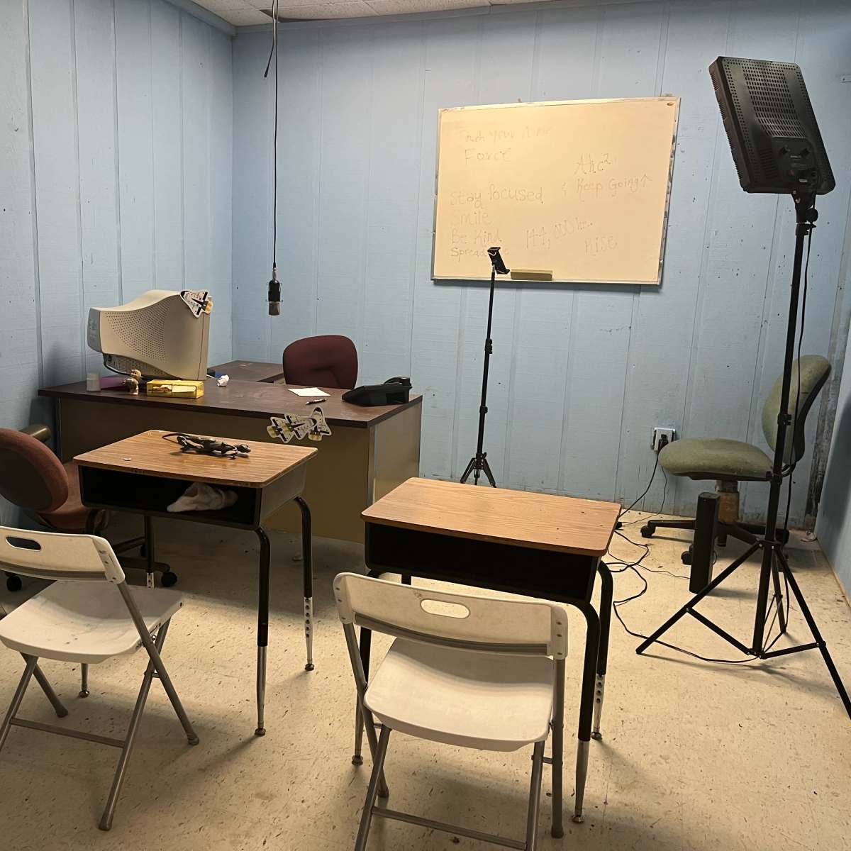 A small, sparsely furnished room with a desk, two empty chairs, and a light stand positioned near a whiteboard on the wall.