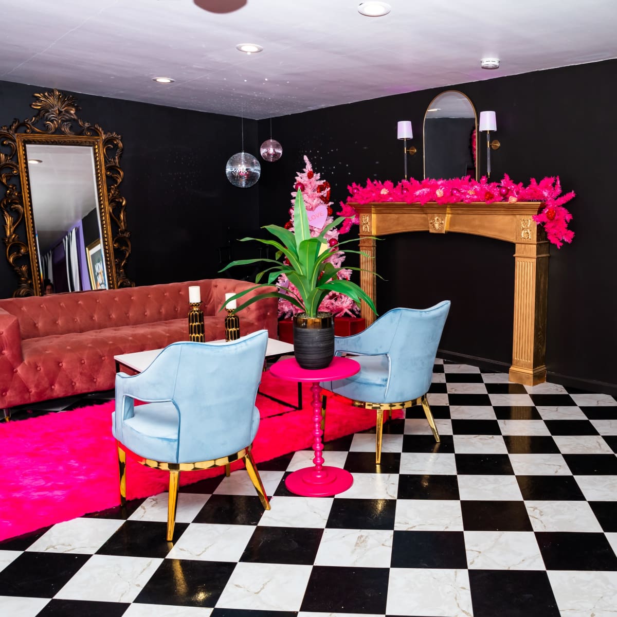 The room features a pink velvet sofa, a black-and-white checkered floor, and a decorative fireplace with vibrant pink accents.
