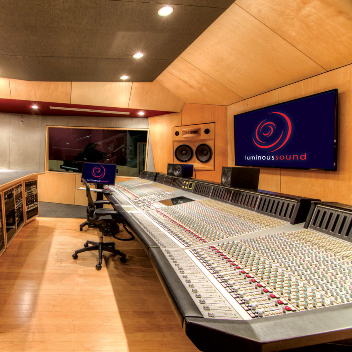 A professional audio recording studio featuring a large mixing console, control desk, and wall-mounted screens.