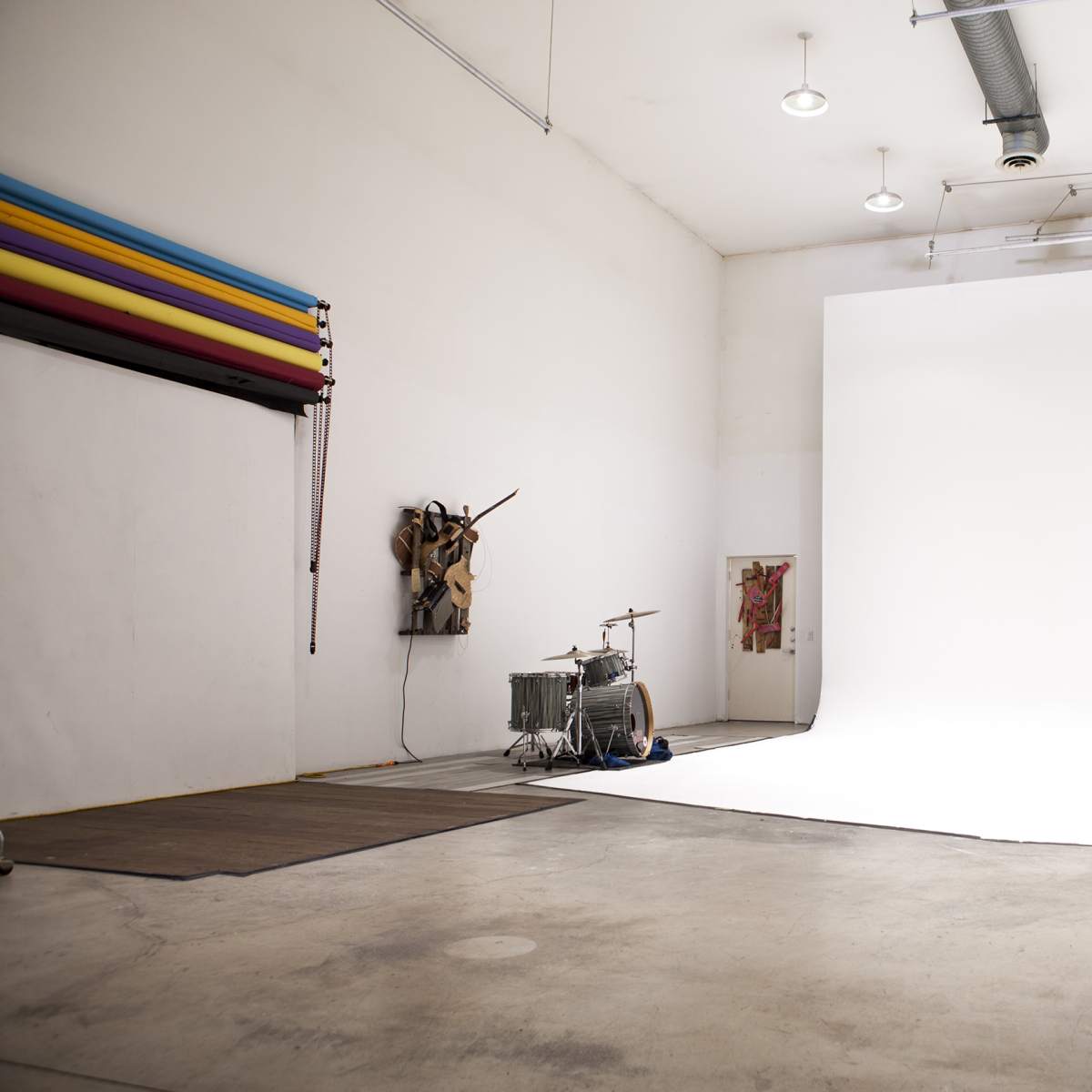 An empty studio space with a white backdrop, a drum set in the corner, and colorful fabric hanging on one wall.