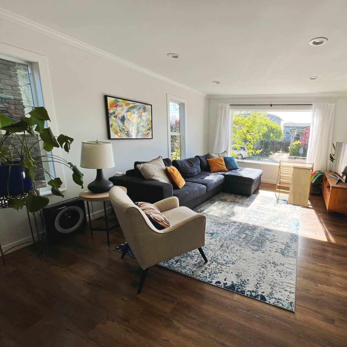 A bright living room features a gray sectional sofa, an armchair, a wooden TV stand, and large windows overlooking a garden.