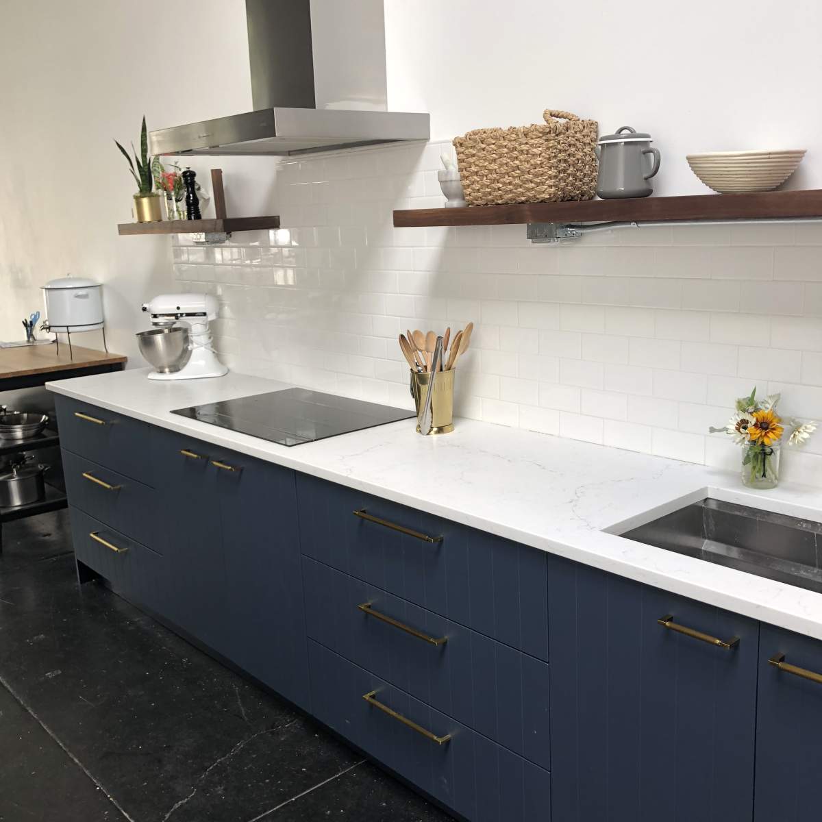 A modern kitchen features dark blue cabinetry, a marble countertop, and stainless steel appliances, with utensils and a small bouquet of flowers on display.
