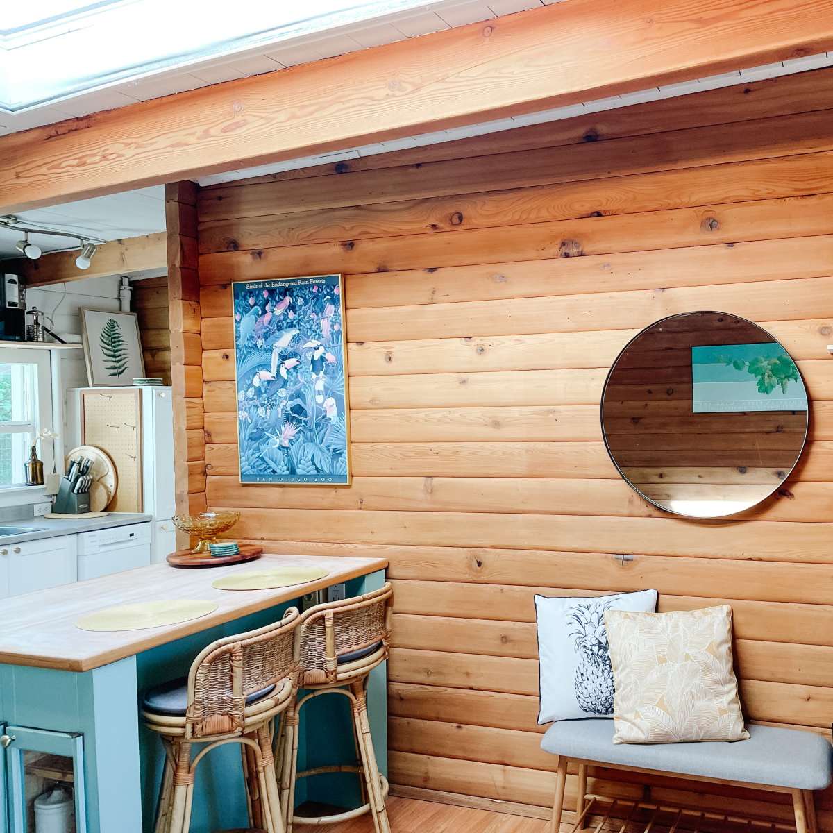 A wooden interior wall, a round mirror, a shelf with decorative items, and a small seating area with cushions beneath a large window.
