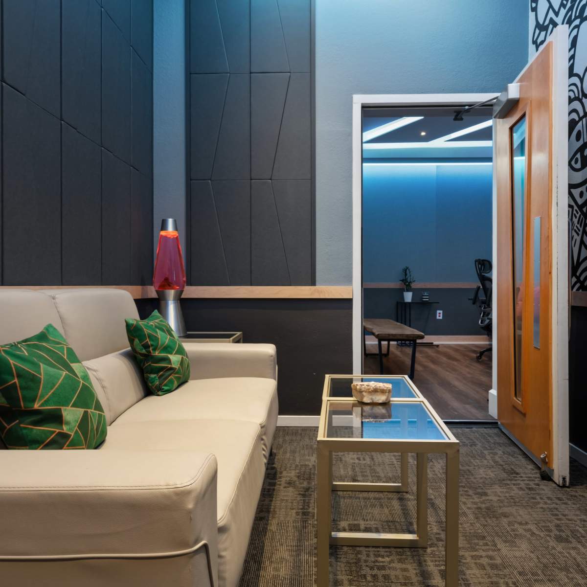 A modern lounge area features a white sofa with decorative green pillows, a glass coffee table, and a doorway leading to a separate room with additional seating.