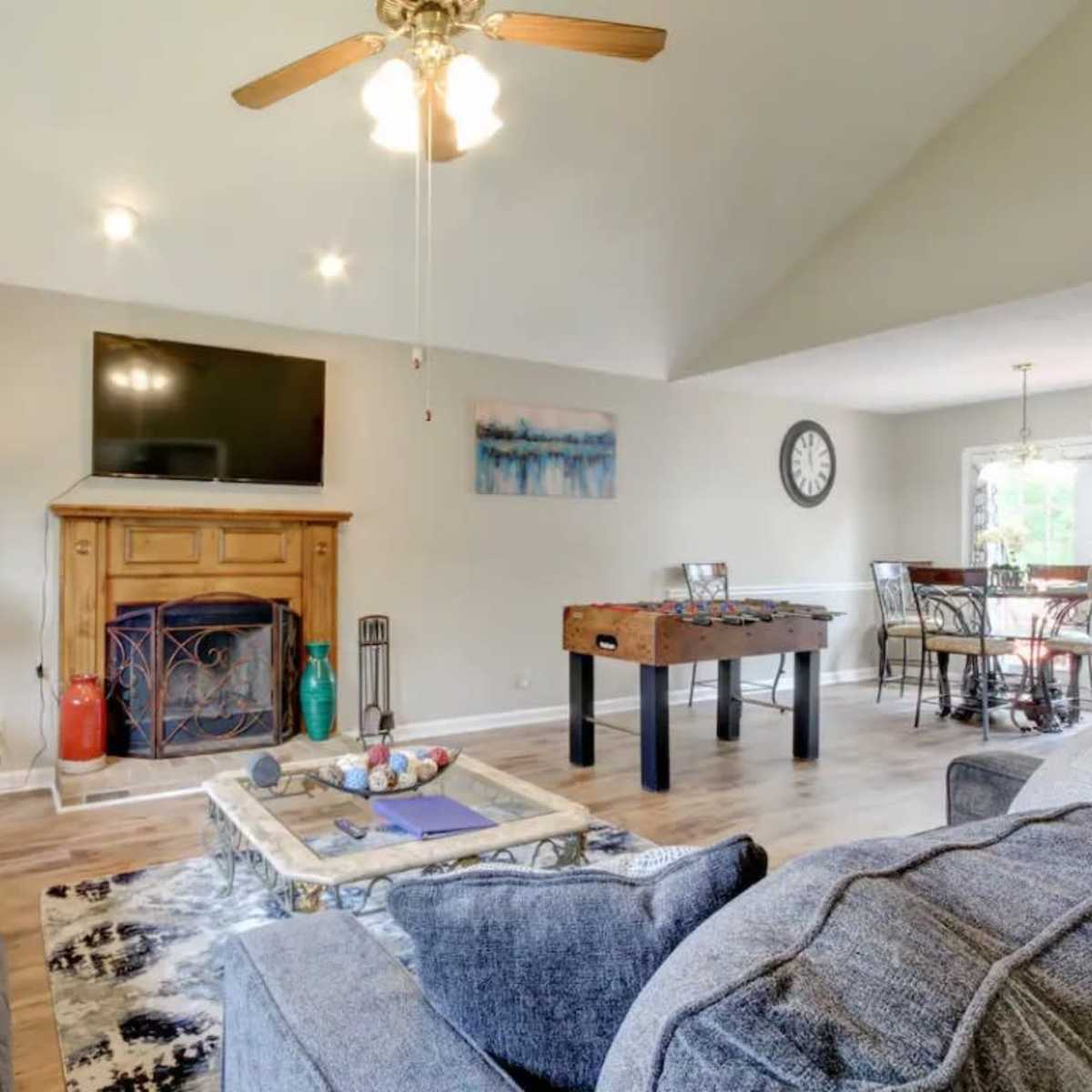 A living room with a sofa, a coffee table featuring decorative items, a fireplace beneath a wall-mounted television, and a foosball table nearby, with a dining area visible in the background.