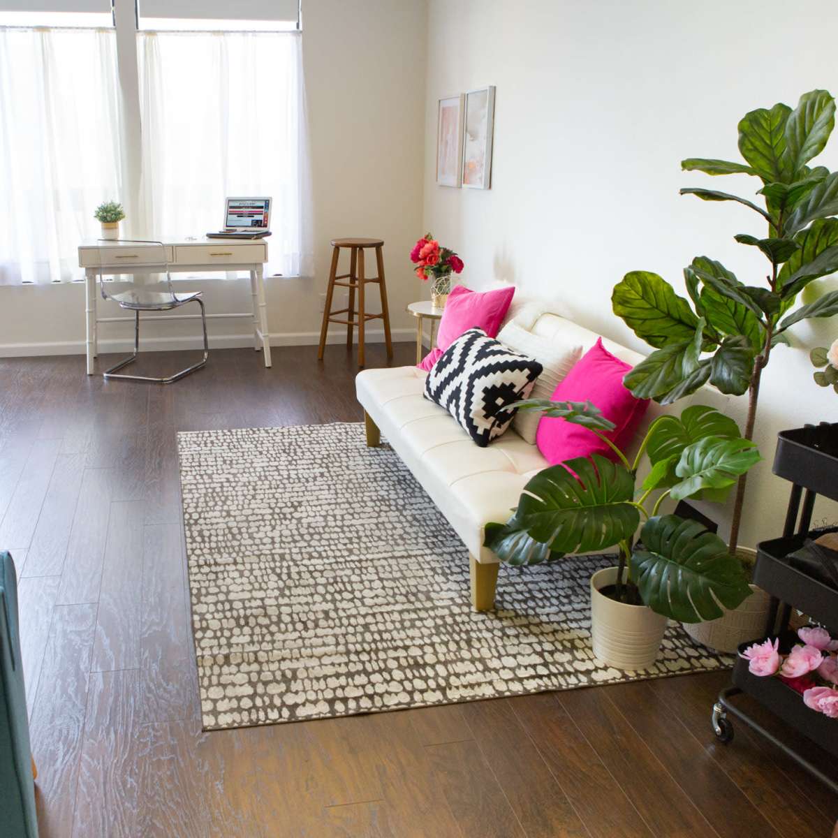 A bright living room featuring a white sofa with pink cushions, a desk, a small table, and decorative plants on a textured rug.